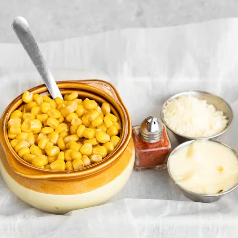 A bowl of corn with a spoon, a shaker of chili powder, and small cups of cheese and mayo.